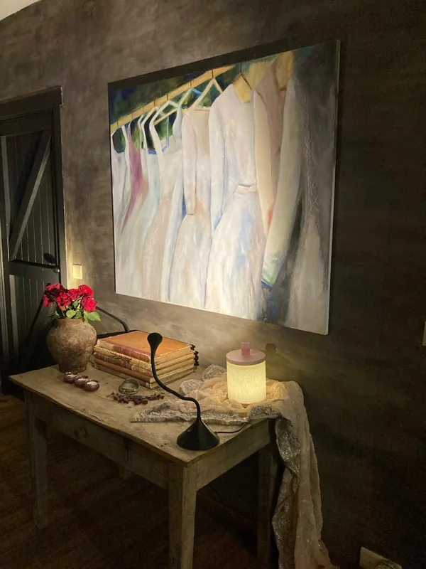 Het schilderij met de jurken boven een tafeltje met een lampje en rode bloemen.